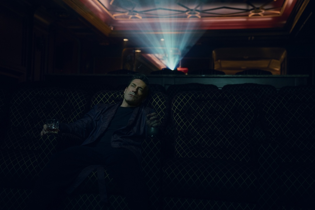 Jon Hamm, as Coop, watches a movie alone in a theater, holding a drink in a whiskey glass in his right hand, and a cigar or cigarette in his left hand.