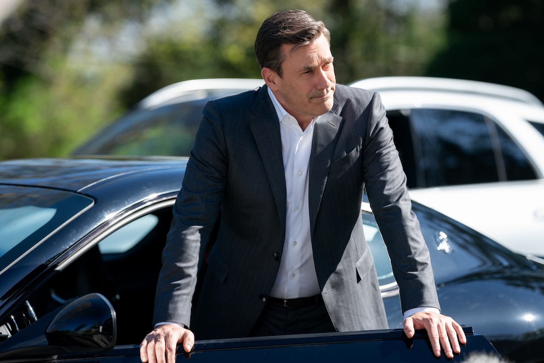 Jon Hamm, as Coop, stands facing the camera, frowning and lips pursed, leaning on the open door of his Maserati.