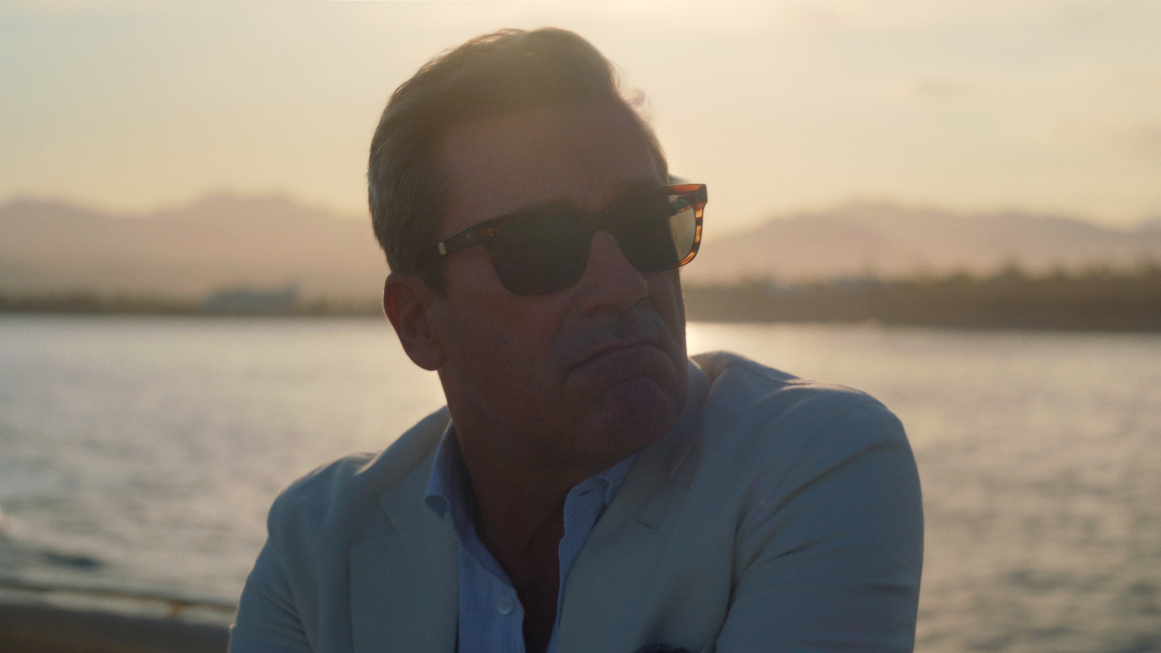 A close-up of Jon Hamm, as Coop, next to a body of water, looking in the distance in his sunglasses