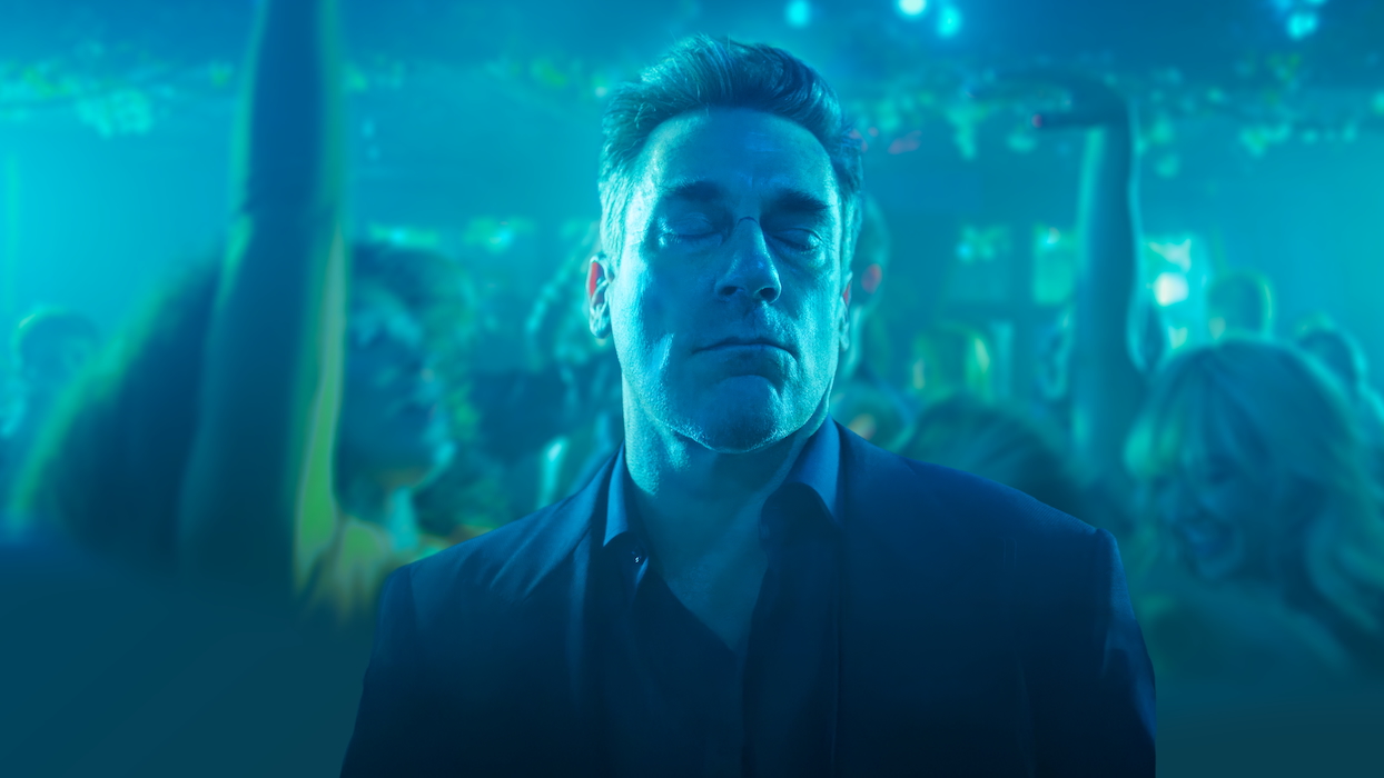 Jon Hamm, as Coop, standing in a club his eyes closed, with people dancing in the background