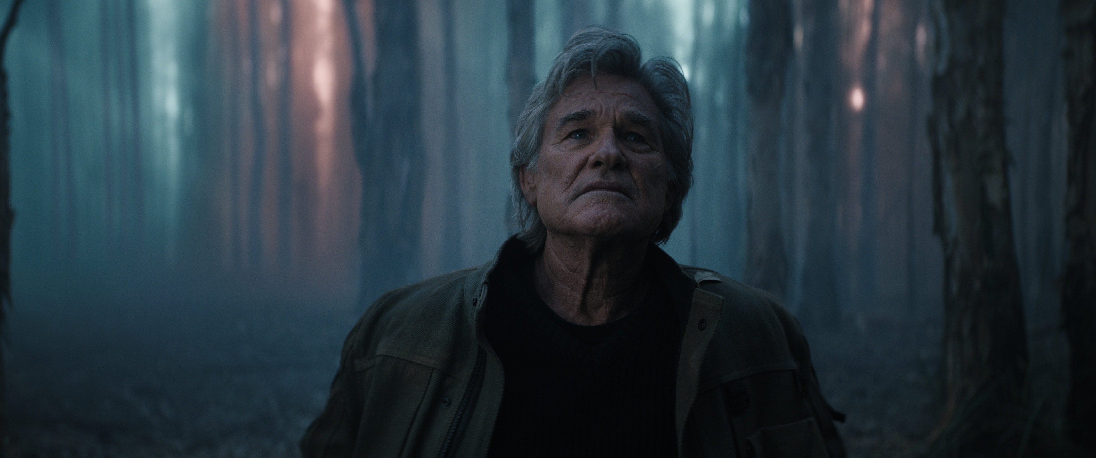Kurt Russell, as Lee Shaw, stands in a dark, misty forest, looking up with a determined expression.
