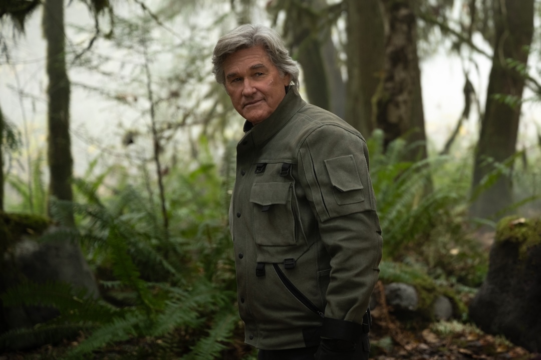 Kurt Russell, as Lee Shaw, wears a green jacket and looks back while standing in a misty, mossy forest.