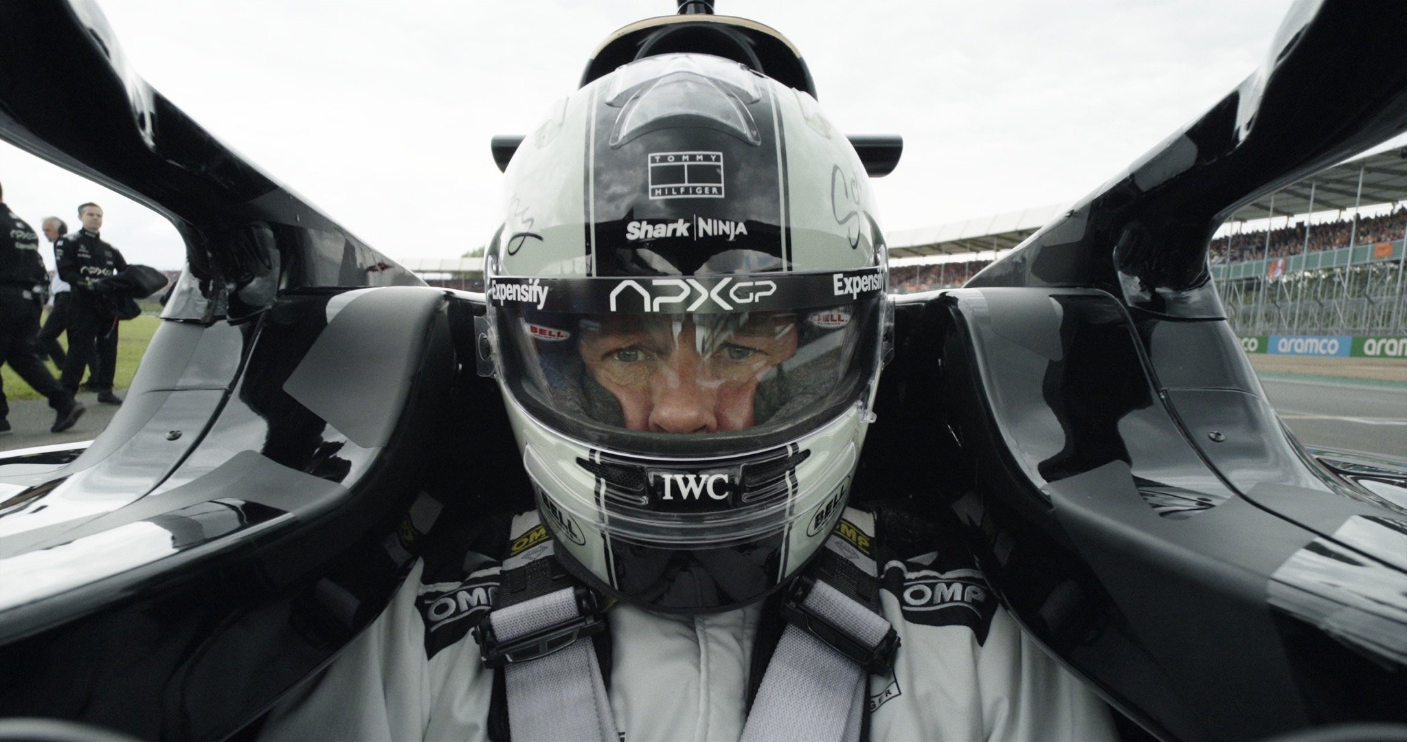A driver-facing onboard view of Brad Pitt as Sonny Hayes in a Formula 1 car, looking focused and wearing an APXGP crash helmet